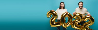 Mavi pankartta 2022 şeklinde altın balonlar tutan beyaz kazaklı mutlu çift.