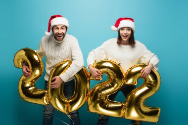 Noel Baba şapkalı heyecanlı çift 2022 model altın balonları mavi üzerinde tutuyorlar.