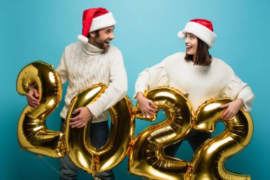 Noel Baba şapkalı şaşkın çift 2022 model mavi balonların yanında birbirlerine bakıyorlar.