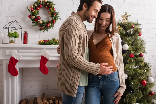 joyful man touching tummy of smiling wife near decorated fireplace and christmas tree