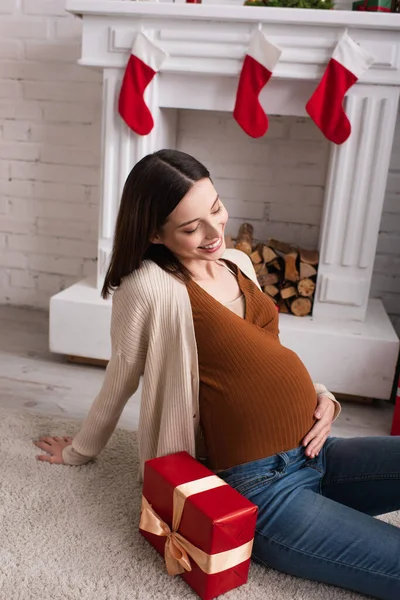 Cheerful Pregnant Woman Touching Tummy While Sitting Gift Box Fireplace — Stock Photo, Image