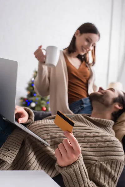 man with laptop holding credit card near blurred wife in living room