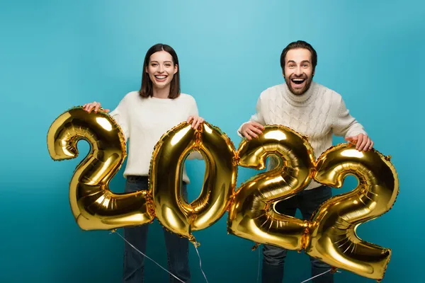 stock image cheerful couple looking at camera while holding golden balloons in shape of 2022 on blue
