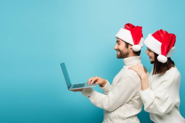 Noel Baba şapkalı ve sıcak kazaklı mutlu çiftin mavi dizüstü bilgisayara bakışları.
