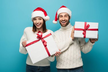 Beyaz kazaklı ve Noel Baba şapkalı neşeli bir çift hediyeler taşıyor ve mavi kameraya bakıyor.