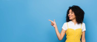 pleased african american woman looking away while pointing with finger on blue, banner clipart