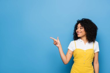 joyful african american woman looking away and pointing with finger on blue clipart