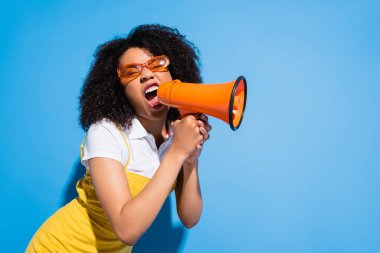 agitated african american woman in trendy eyeglasses screaming in megaphone on blue clipart