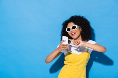 Şık güneş gözlüklü neşeli Afrikalı Amerikalı kadın mavi üzerine cep telefonu kullanıyor.