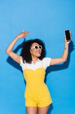 Güneş gözlüklü ve sarı tulumlu neşeli Afro-Amerikalı kadın mavi üzerine cep telefonunu işaret ediyor.