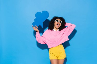 Güneş gözlüklü afro-amerikan kadın boş düşünce baloncuğu tutuyor ve mavi üzerine bağırıyor.