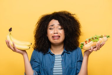 upset african american woman choosing between fresh bananas and hot dog isolated on yellow clipart