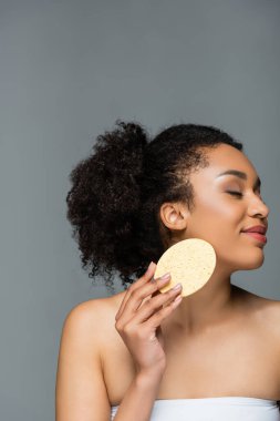 Mutlu Afro-Amerikalı kadın, gözleri kapalı, yüzünü makyaj süngeriyle siliyor, gri renkte.