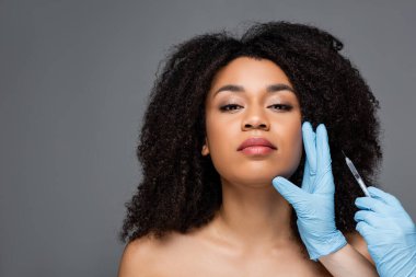 Kauçuk eldivenli kozmetik uzmanı elinde güzellik enjeksiyonuyla, gri üzerinde izole edilmiş Afrikalı Amerikalı bir kadının yüzüne yakın bir yerde.