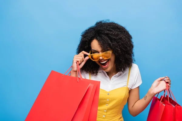 兴奋的非洲裔美国女人 拿着购物袋摸着蓝色的时髦眼镜 — 图库照片