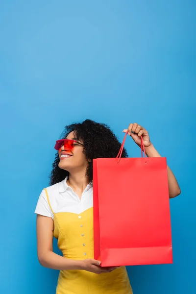 青で隔絶されたサンゴのショッピングバッグを見せる赤い眼鏡のアフリカ系アメリカ人女性 — ストック写真