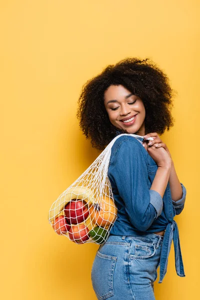 happy african american woman carrying string bag with fresh fruits on yellow
