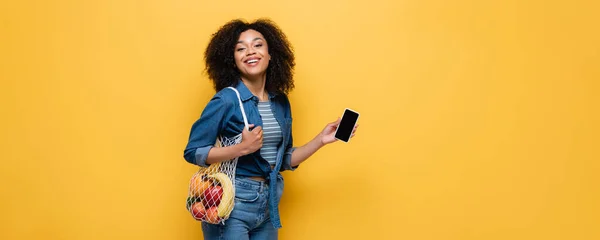 Счастливая Африканская Американка Держит Смартфон Струнный Мешок Фруктами Изолированы Желтый — стоковое фото