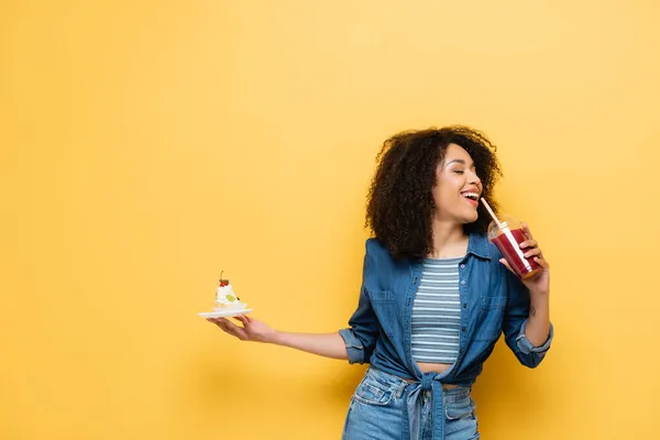 Happy African American Woman Holding Cupcake While Drinking Fresh Smoothie — Stock Photo, Image