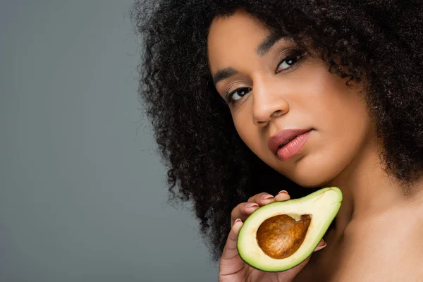 young african american woman with natural makeup holding half of fresh avocado isolated on grey