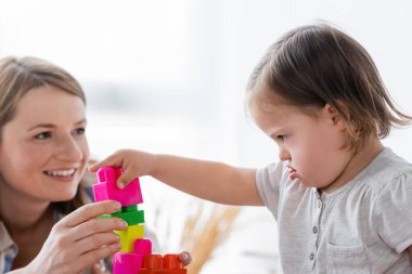 Concentrated kid with down syndrome playing building blocks near blurred smiling mother at home  clipart