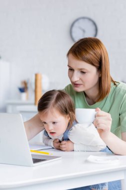 Down sendromlu ve dizüstü bilgisayarı mutfakta olan bir kızın bardağını tutan kadın. 