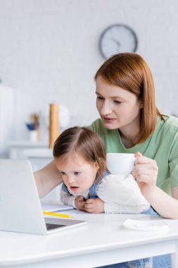 Woman holding cup near daughter with down syndrome and laptop in kitchen  clipart