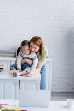Smiling woman pointing at laptop near daughter with down syndrome in kitchen  clipart