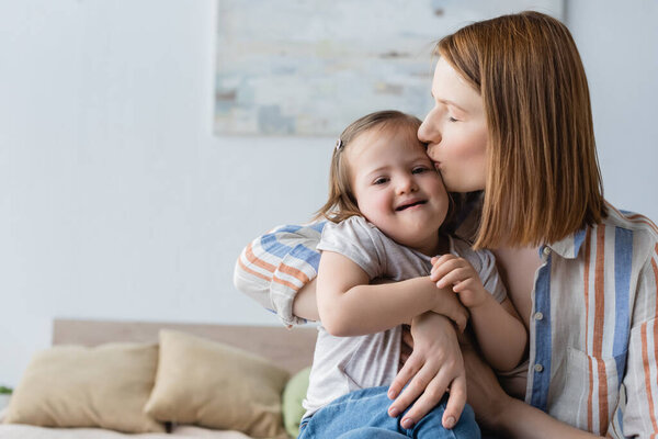Мать целует улыбающуюся дочку с синдромом Дауна в спальне 