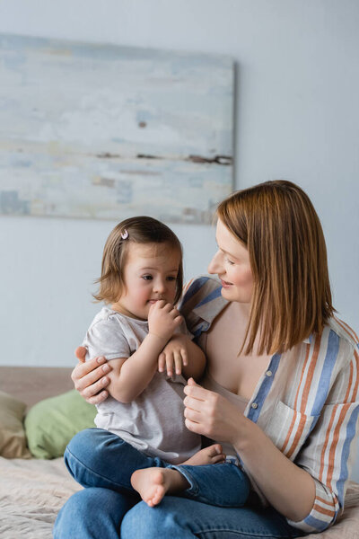 Позитивная женщина смотрит на дочь малыша с синдромом Дауна в спальне 