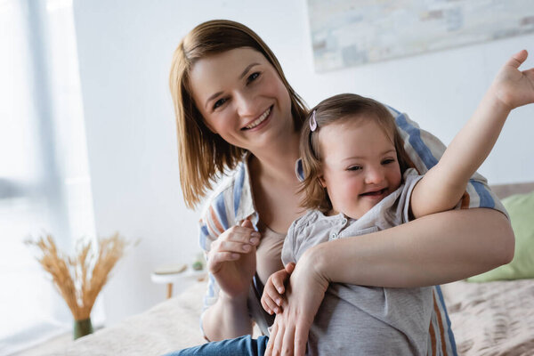 Улыбающаяся женщина смотрит в камеру, обнимая дочь с синдромом Дауна на кровати 