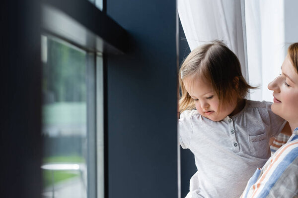 Woman and toddler girl with down syndrome standing near window at home 