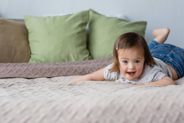Веселый ребенок с синдромом Дауна смотрит в камеру на кровати 