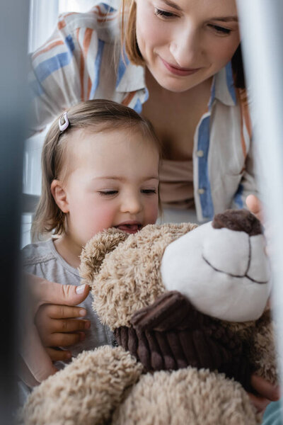 Веселый ребенок с синдромом Дауна, смотрящий на плюшевого мишку рядом с матерью дома 