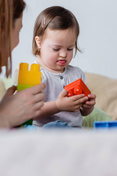 Toddler girl with down syndrome holding building block near mother at home 