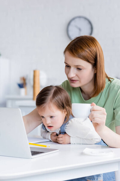 Woman holding cup near daughter with down syndrome and laptop in kitchen 