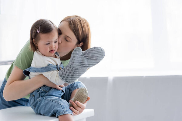 Родитель целует ребенка с синдромом Дауна держа выпечку перчатку на кухне 