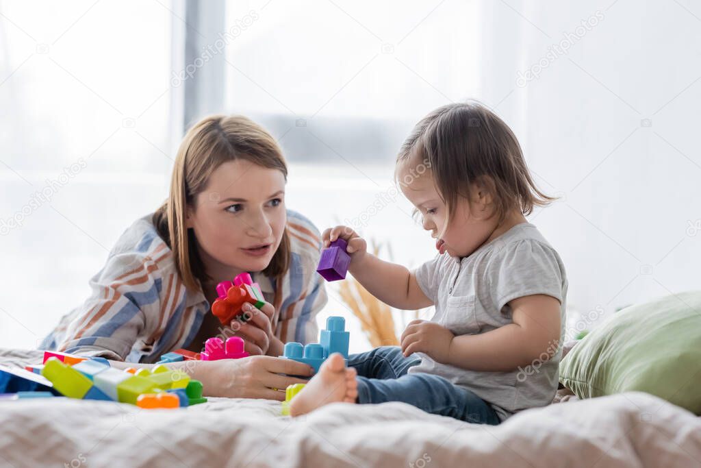 Child with down syndrome playing building blocks near mom in bedroom 