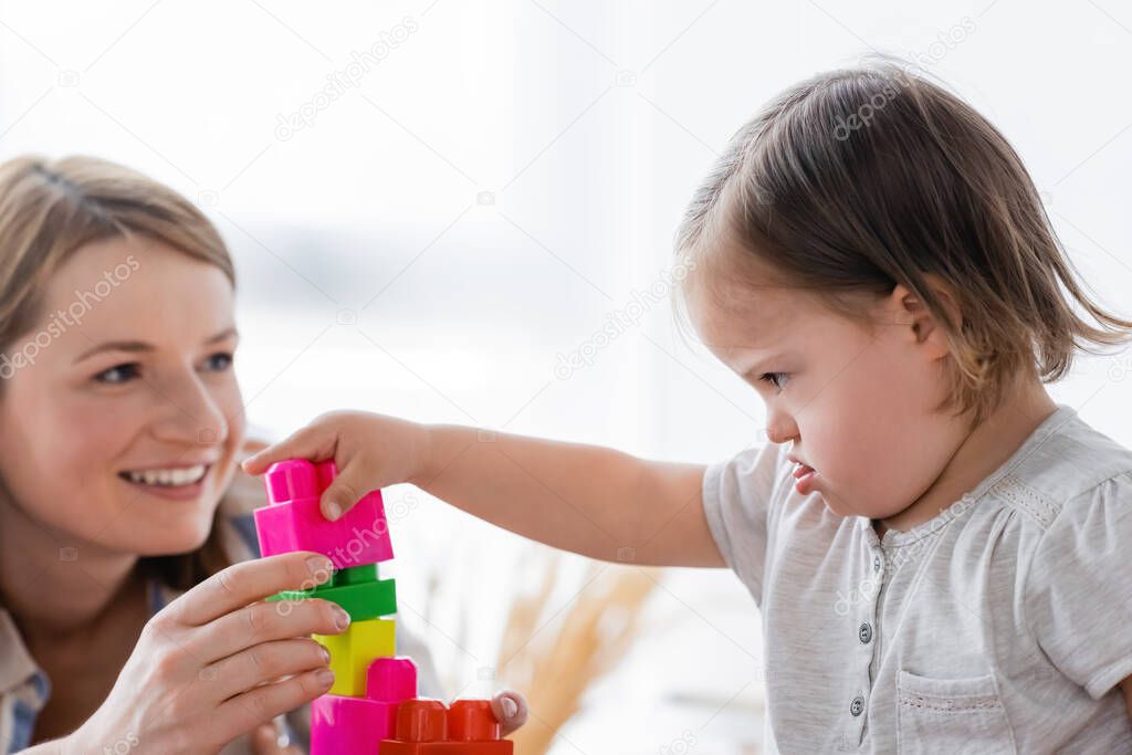 Concentrated kid with down syndrome playing building blocks near blurred smiling mother at home 