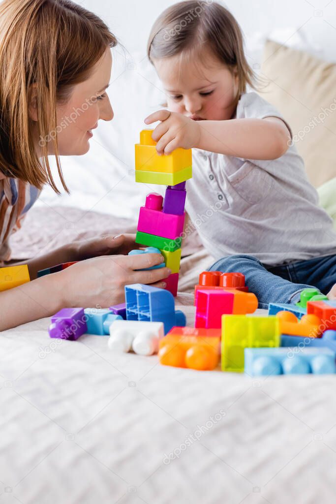 Happy woman looking at toddler child playing building blocks on bed 