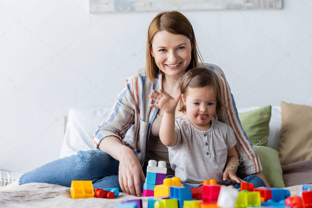 Woman and child with down syndrome waving hand at camera near building blocks on bed 