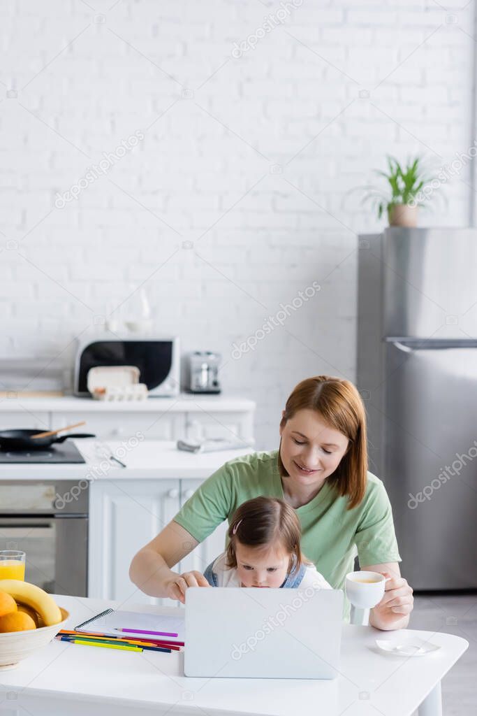 Smiling woman holding coffee near laptop and daughter with down syndrome in kitchen 