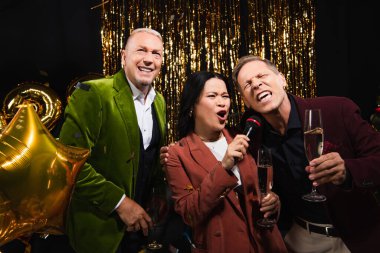 Siyah arka planda parti sırasında şampanyalı Asyalı kadın olgun arkadaşlarıyla karaoke yapıyor. 