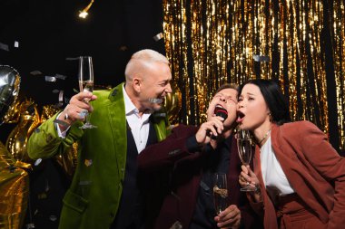 Şampanyalı çok ırklı arkadaşlar konfeti ve siyah arka planda süslemeler eşliğinde karaoke yapıyorlar. 