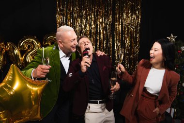 Yeni yıl partisinde arkadaşlarının yanında elinde şampanya tutan Asyalı kadın siyah arka planda karaoke yapıyor. 