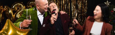 Arkadaşlarının yanında şampanya tutan neşeli Asyalı kadın siyah arka plandaki Noel partisinde karaoke söylüyor. 