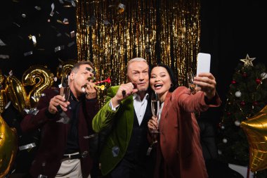 Şampanyalı gülümseyen Asyalı kadın, mikrofonu olan ve siyah arka planda parti boynuzları olan arkadaşlarıyla selfie çekiyor. 
