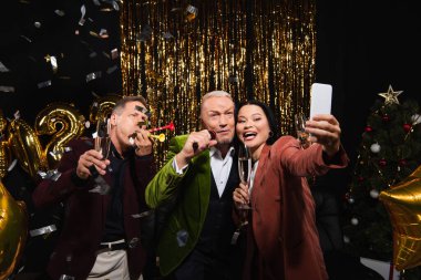 Smiling asian woman with champagne taking selfie with friends with microphone and party horns on black background  clipart