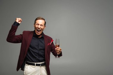 Excited mature man with glass of champagne showing yes gesture isolated on grey  clipart
