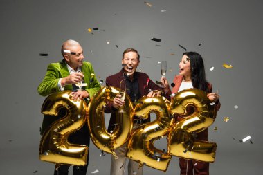 Şampanyalı heyecanlı ırklar arası arkadaşlar 2022 numaralı balonların yanında duruyorlar ve gri arkaplanda konfeti var. 